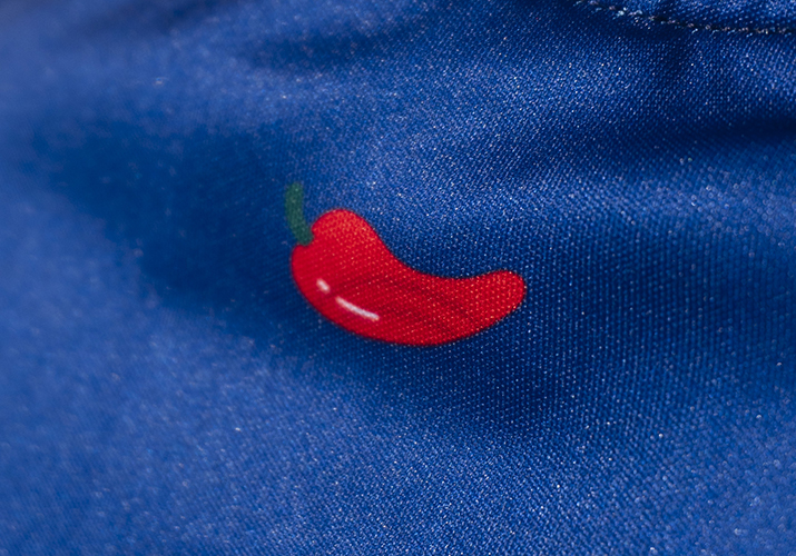 Fotografía de bordado en camiseta Montdebó | Carlos Sainz Primer plano de un pequeño chile rojo bordado sobre el tejido azul de una camiseta Montdebó.