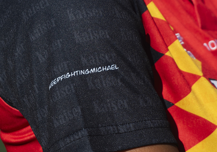 Detalle de camiseta Montdebó | Michael Schumacher Primer plano de la manga negra de una camiseta Montdebó con la inscripción «Keep Fighting Michael».
